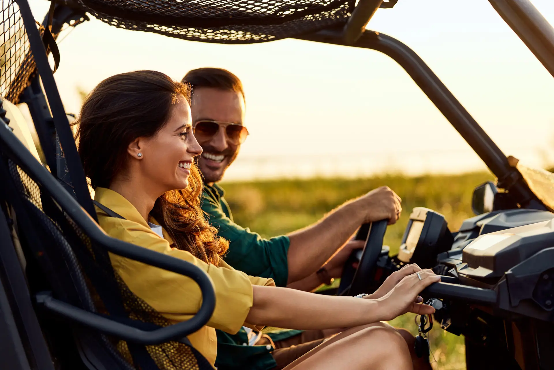 Une femme et un homme souriants se promènent dans un véhicule tout-terrain ouvert au coucher du soleil. La femme est sur le siège passager et l'homme, qui porte des lunettes de soleil, conduit. L'herbe verte et la lumière chaude du soleil sont visibles à l'arrière-plan.