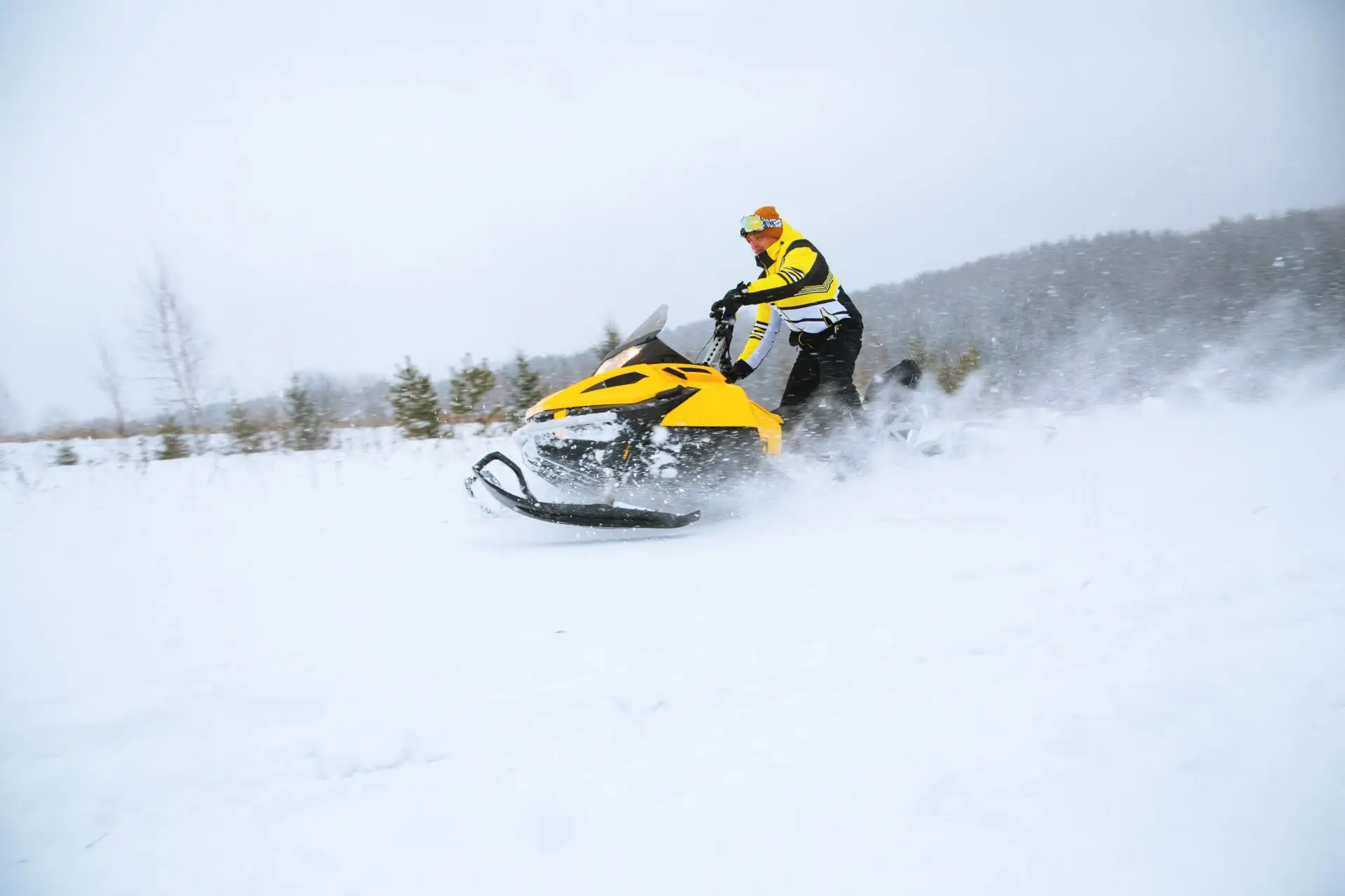 Une personne portant une veste et un casque jaune et noir conduit une motoneige jaune à grande vitesse dans un paysage enneigé avec des arbres et des collines en arrière-plan. La neige est projetée derrière la motoneige.