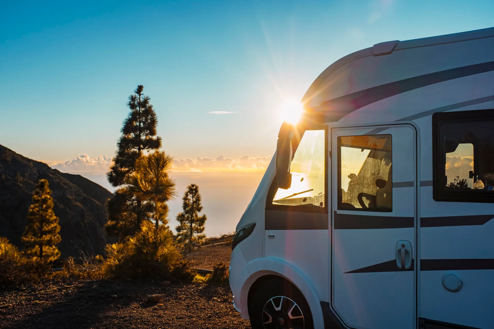 Un camping-car blanc garé au sommet d'une colline au coucher du soleil, la lumière du soleil traversant les pins et se reflétant sur l'océan en arrière-plan.