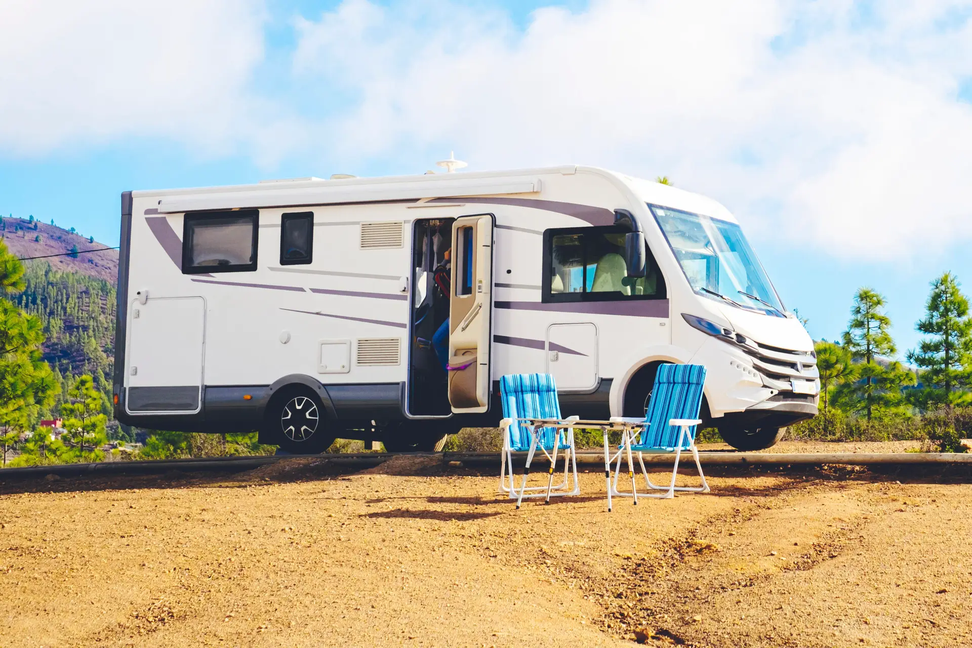 Un camping-car blanc est garé sur une aire en terre battue, porte ouverte. Deux chaises pliantes bleues et blanches et une petite table sont installées à l'extérieur. Des arbres et des collines sont visibles à l'arrière-plan sous un ciel partiellement nuageux.