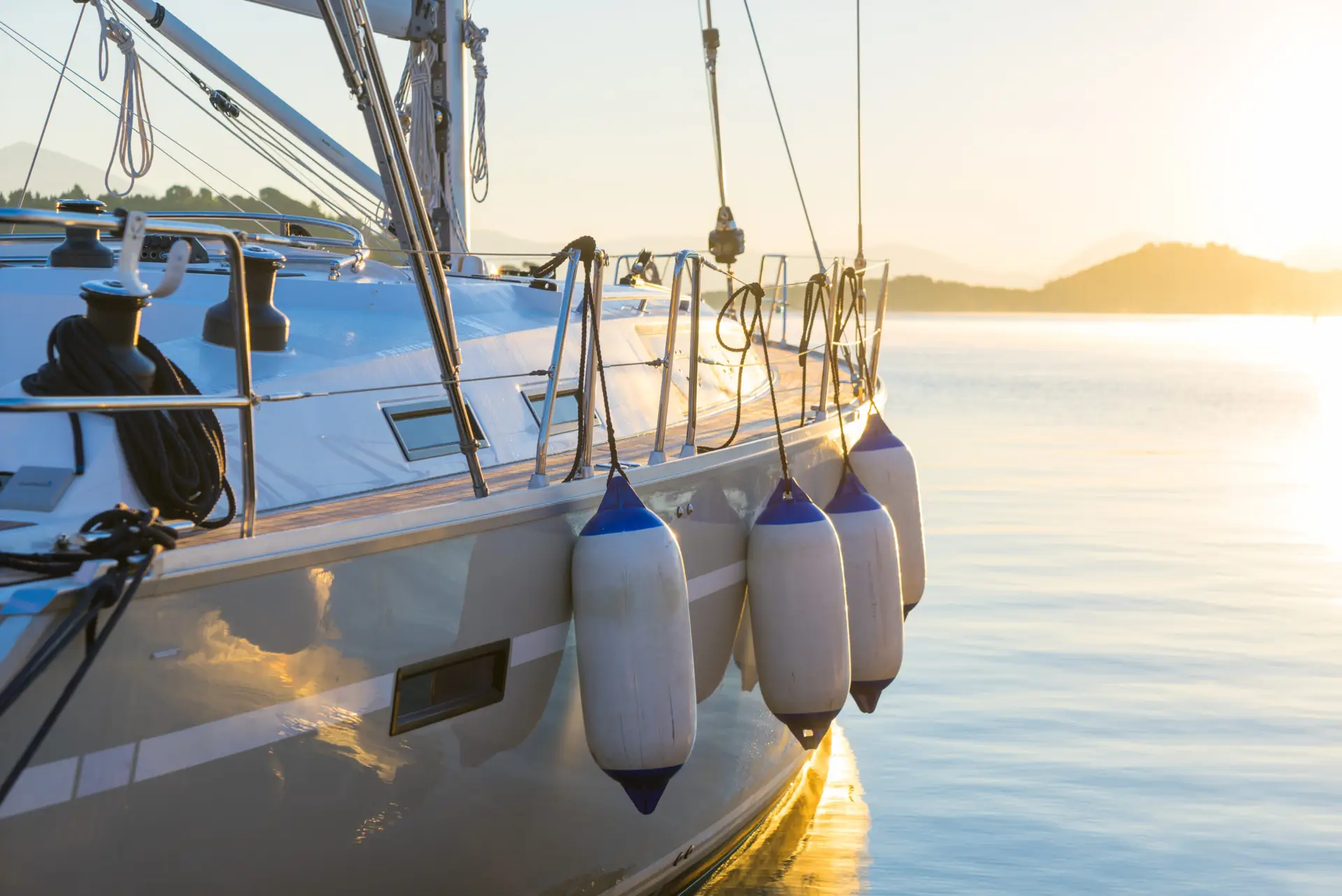 Gros plan sur le flanc d'un voilier au lever ou au coucher du soleil, la lumière du soleil se reflétant sur l'eau. Des défenses bleues et blanches sont suspendues le long du bord du bateau, et l'on peut voir à l'arrière-plan des eaux calmes et des collines lointaines.