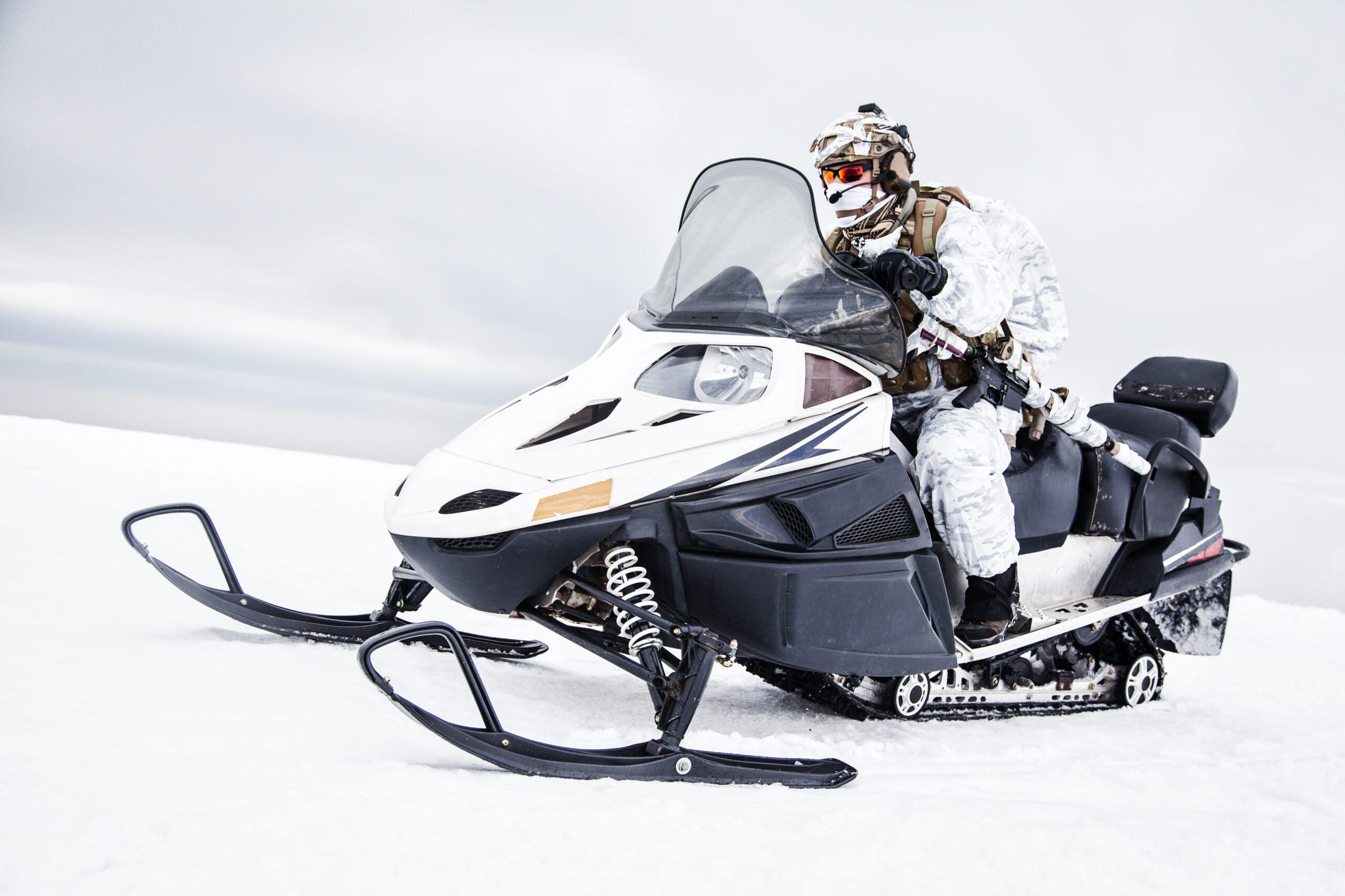 Une personne portant une tenue de camouflage hivernal et des lunettes de protection conduit une motoneige noire et blanche dans un paysage enneigé sous un ciel nuageux.