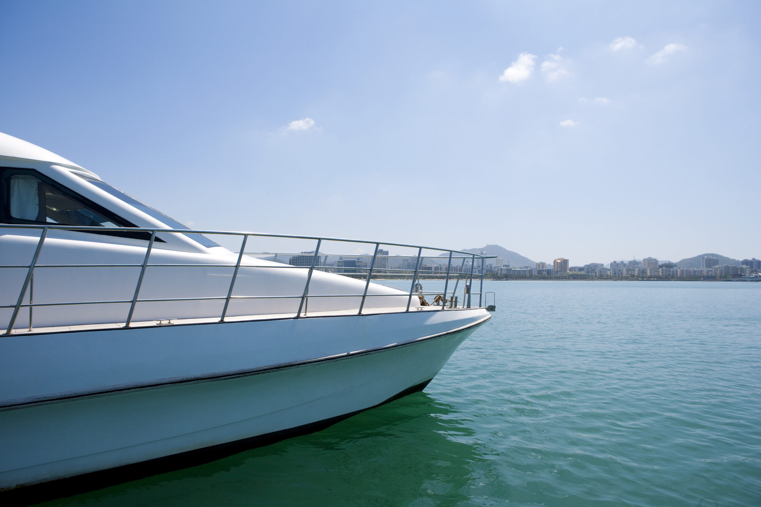 Vue de face d'un yacht blanc flottant sur une eau calme et turquoise, avec en arrière-plan la ligne d'horizon d'une ville et des collines visibles sous un ciel bleu clair.